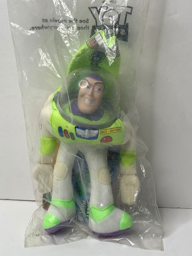 May include: A green and white plush toy of Buzz Lightyear from the Toy Story movie. The toy is in a plastic bag with the words "Toy Story" printed on it. The toy has a purple helmet with a clear visor and a purple shirt. The toy is holding a green laser gun.