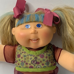 Może przedstawiać: Lalka Cabbage Patch Kid z blond włosami i niebieskimi oczami. Lalka ma na sobie zielono-brązowy kwiatowy top, bordową koszulę i niebieskie dżinsy. Lalka ma różowe kokardki we włosach.