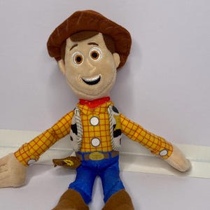 May include: A plush toy of a cartoon cowboy character. The toy has a brown hat, brown hair, and a smiling face. He wears a yellow plaid shirt, blue pants, brown boots, and a red neckerchief. The toy has a white vest with black spots.