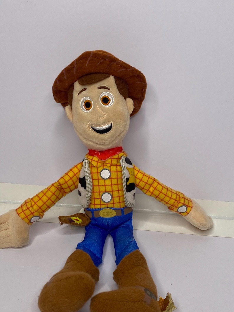 May include: A plush toy of a cartoon cowboy character. The toy has a brown hat, brown hair, and a smiling face. He wears a yellow plaid shirt, blue pants, brown boots, and a red neckerchief. The toy has a white vest with black spots.