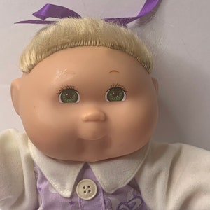 Könnte beinhalten: Eine Cabbage Patch Kid Puppe mit blonden Haaren, grünen Augen und einer lila Schleife. Die Puppe trägt ein lila-weißes Kleid mit einem weißen Kragen und Knöpfen.