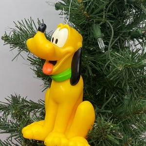 Pode incluir: Um ornamento do cão Pluto em amarelo com nariz preto, orelhas pretas e uma coleira verde brilhante. A personagem tem uma expressão de boca aberta com uma língua laranja visível. Um gancho de metal prateado está preso ao topo da cabeça.