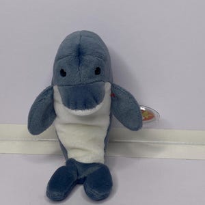 Puede incluir: Un peluche de delfín en tonos azules y blancos. El delfín tiene una cabeza redondeada, ojos negros pequeños y brazos con forma de aleta. Una pequeña etiqueta está adherida al delfín. El juguete está diseñado para parecerse a un delfín.