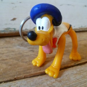 Puede incluir: Un llavero de plástico amarillo con un perro de dibujos animados que lleva un sombrero azul. El perro tiene la lengua larga que sobresale y está de pie sobre sus cuatro patas.