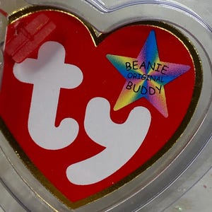 K&ouml;nnte beinhalten: Ein rotes herzf&ouml;rmiges Etikett mit goldenem Rand und den wei&szlig;en Buchstaben "ty". Ein sternf&ouml;rmiges Emblem mit den Worten "Beanie Original Buddy" befindet sich auf der rechten Seite. Das Etikett ist in einem transparenten Kunststoffherz eingeschlossen.