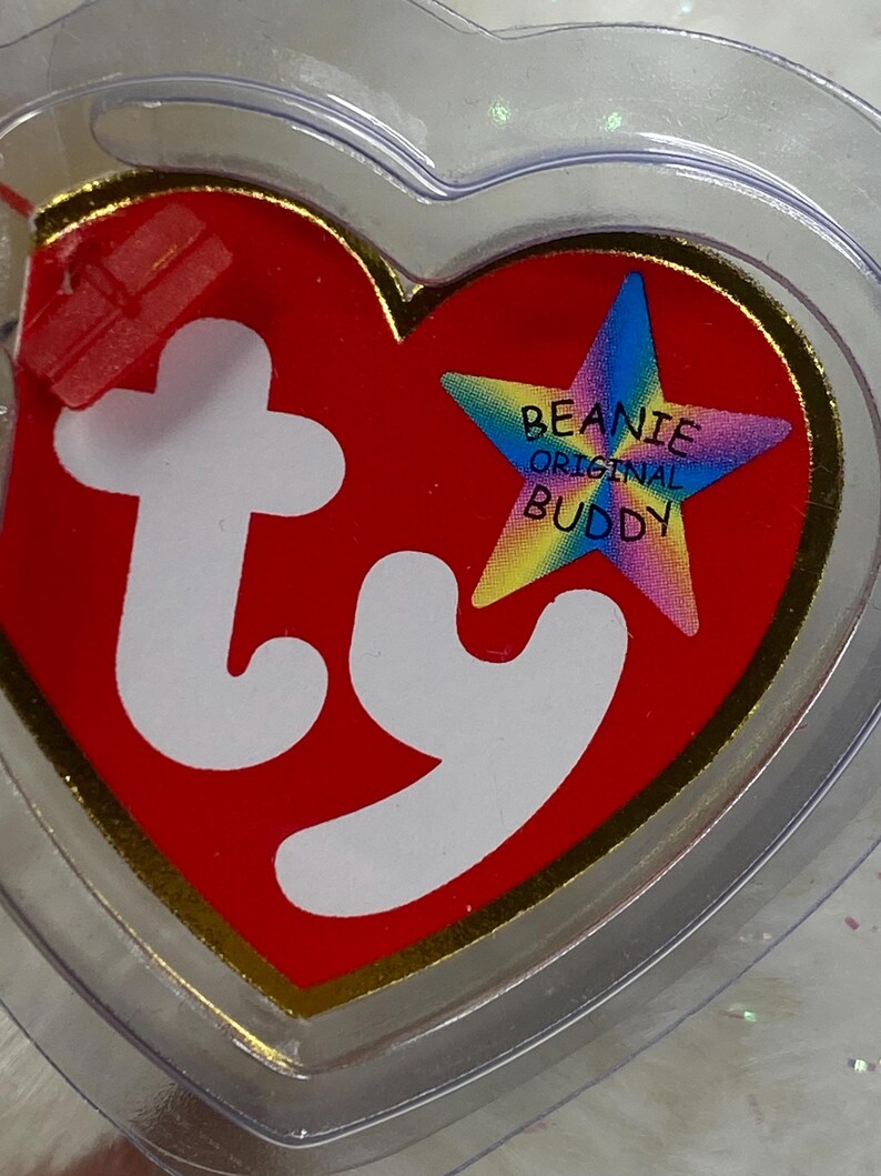 K&ouml;nnte beinhalten: Ein rotes herzf&ouml;rmiges Etikett mit goldenem Rand und den wei&szlig;en Buchstaben "ty". Ein sternf&ouml;rmiges Emblem mit den Worten "Beanie Original Buddy" befindet sich auf der rechten Seite. Das Etikett ist in einem transparenten Kunststoffherz eingeschlossen.