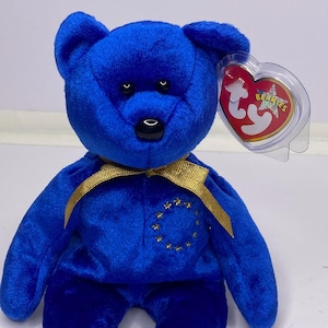 May include: A blue plush bear with a gold ribbon tied around its neck. The bear has a gold embroidered design on its chest and a red heart-shaped tag that reads "Beanie Babies".