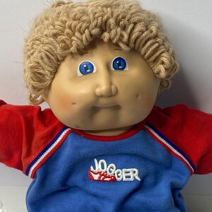 May include: A Cabbage Patch Kid doll with blonde yarn hair wearing a blue and red jogging outfit with the word "Jogger" embroidered on the front.