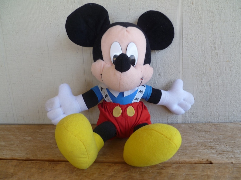 Może przedstawiać: Pluszowa zabawka Mickey Mouse w niebiesko-białej pasiastych koszuli, czerwonych ogrodniczkach i ż&oacute;łtych butach. Zabawka siedzi na drewnianej powierzchni.
