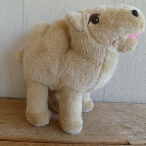 May include: A plush camel toy with a light brown fur, standing on a wooden surface. The camel has a brown eye and a pink mouth.