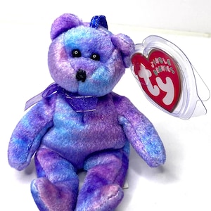 May include: A plush teddy bear in shades of purple and blue, with a matching bow and ribbon. The bear has black eyes and a red heart-shaped tag attached to its ear. The tag reads "Jingle Beanies" and "ty".