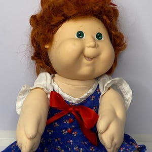 May include: A vintage doll with a round face, green eyes, and red curly hair. The doll wears a blue dress with a floral pattern and a red bow. A red bow is also in the doll's hair. The doll's arms and hands are visible.