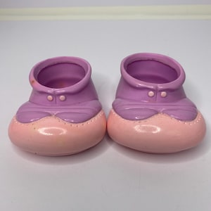 May include: A pair of small, plastic baby shoes. The shoes are two-toned, with a light purple upper and a pink lower section. Each shoe has two small, round buttons and a decorative seam.