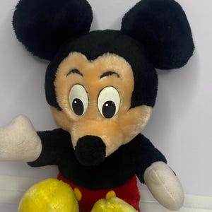 May include: A plush Mickey Mouse doll with black ears, a black head, and a tan face. The doll has white gloves, red shorts, and yellow shoes. The doll is a classic Disney character.