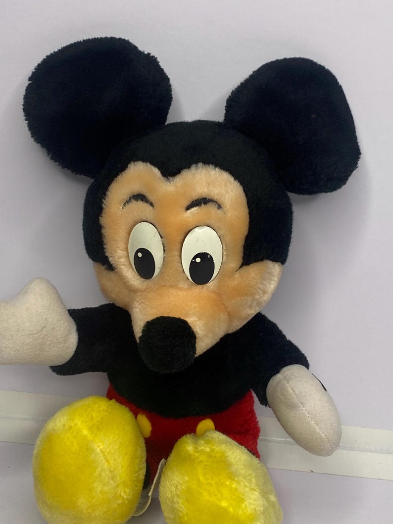 May include: A plush Mickey Mouse doll with black ears, a black head, and a tan face. The doll has white gloves, red shorts, and yellow shoes. The doll is a classic Disney character.