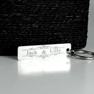 May include: A rectangular silver keychain with the names "Jack & Lilly" engraved in a decorative font. The keychain has a silver key ring attached. A black woven basket is in the background.