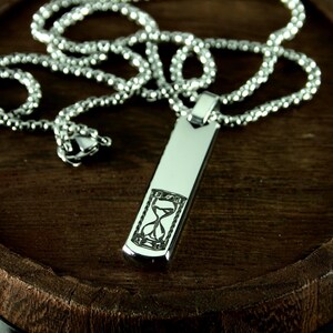 May include: A silver-colored pendant necklace with a rectangular pendant featuring an engraved hourglass design. The chain is made of small, round, interconnected links. The necklace rests on a dark brown, wooden surface.