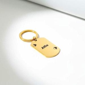 Puede incluir: Llavero rectangular dorado con un anillo redondo. El llavero tiene esquinas redondeadas y presenta el nombre "Alfie" en texto negro, junto con símbolos de picas. Un accesorio personalizado.