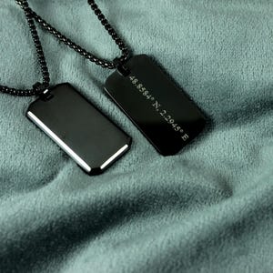 May include: Two black dog tag necklaces with matching chains on teal fabric. One tag is blank, the other has engraved coordinates: 48.8584° N, 2.2945° E. The tags have a shiny, reflective surface.