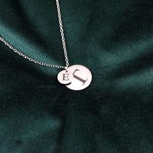 May include: A silver necklace with two circular pendants. The larger pendant has the letter "J" engraved, and the smaller pendant has the letter "E". The necklace chain is also silver.