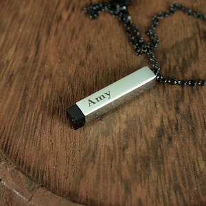 Puede incluir: Collar con colgante rectangular plateado grabado con el nombre "Amy". El colgante tiene un extremo negro y cuelga de una cadena oscura. El collar descansa sobre una superficie de madera.
