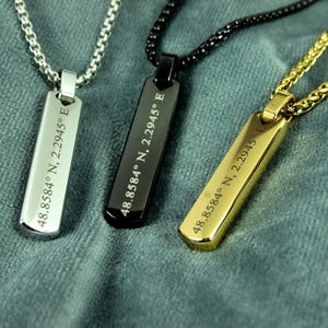 Puede incluir: Tres collares con colgantes rectangulares y cadenas. Las etiquetas son plateadas, negras y doradas, cada una grabada con "48.8584° N, 2.2945° E". Los collares descansan sobre una superficie texturizada, verde azulado.