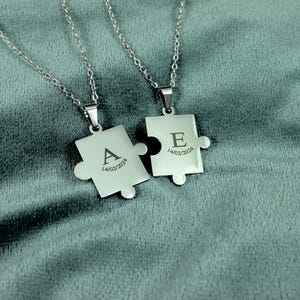 May include: Two silver puzzle piece pendants, each engraved with a letter and date: "A" and "E", "14/02/2024". The necklaces hang on fine chains. The background is a soft, grey-green fabric.