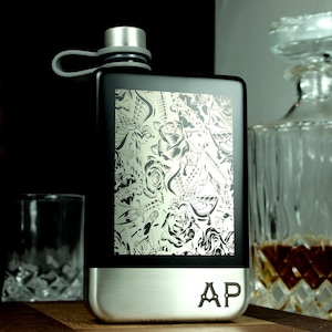 May include: Black flask with a silver base and a decorative floral and butterfly design. The flask has a silver cap and a grey loop. The initials "AP" are engraved on the base. A glass decanter and a wooden barrel are in the background.