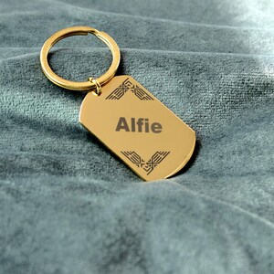 May include: Gold-colored rectangular keychain with the name "Alfie" engraved in the center. The keychain has decorative corner designs and a gold ring. The background is a textured, teal-colored fabric.