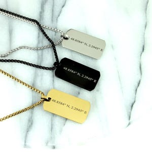 May include: Three dog tag necklaces in silver, black, and gold. Each tag is rectangular with rounded corners and features the engraved coordinates "48.8584° N, 2.2945° E". The chains are a matching colour to the tags.
