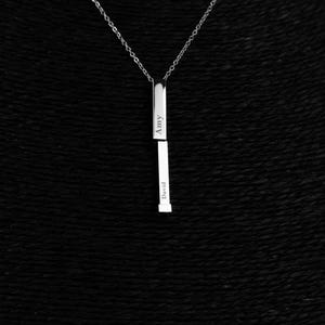 Puede incluir: Un collar con colgante plateado con un diseño de barra vertical. La barra está dividida en dos secciones, con los nombres "Amy" y "David" grabados. El collar cuelga de una delicada cadena sobre un fondo negro.