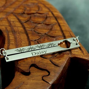 May include: A silver bar necklace with the name "Daisy" engraved on it. The necklace features a heart-shaped cutout and a decorative line of thorns. The necklace is displayed on a carved wooden surface.