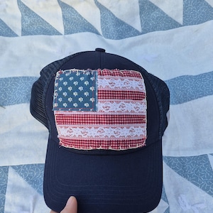 May include: Navy blue trucker hat with a patch featuring a patchwork American flag design. The flag incorporates blue fabric with white stars, red and white checkered fabric, and white lace trim. The hat has a mesh back.