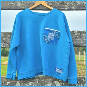 May include: A blue sweatshirt with a crew neck and long sleeves. The sweatshirt features a pocket with a floral pattern and an Ace of Hearts playing card design. A small rectangular patch with the text "ACE + ACE" is sewn on the lower right side.