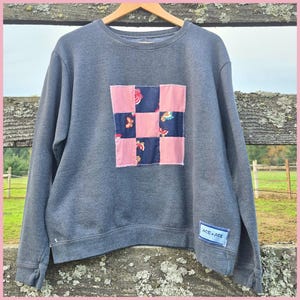 May include: A gray sweatshirt with a patchwork design on the front. The patchwork is a square of pink and navy blue squares. The navy blue squares have a floral pattern. The sweatshirt has the text "ACE + ACE" on the sleeve.