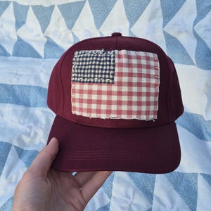 May include: A burgundy baseball cap with a sewn-on patch resembling a flag. The flag design features a navy blue and white checkered square in the upper left corner and a red and white checkered pattern on the right. The cap has a curved brim.