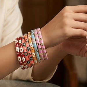 Puede incluir: Un conjunto de siete brazaletes finos de metal que se llevan en la muñeca, con un degradado de colores que incluye rojo, naranja, amarillo, azul claro y rosa. Cada brazalete está decorado con un patrón repetitivo de pequeñas flores blancas.