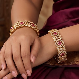 May include: Two gold-toned bangles featuring a repeating floral-inspired pattern set with small clear stones and a central square-cut pink stone. The bangles are worn on the wrists, showcasing a detailed metalwork design with a polished finish.