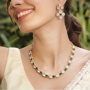 May include: A gold-toned necklace and matching drop earrings set featuring alternating clear and emerald green circular stones. The necklace consists of a single strand of round gems, while the earrings display a cluster of stones in a floral shape.