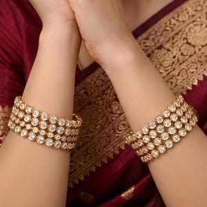 Pode incluir: Pulseiras douradas com várias fileiras de pedras preciosas redondas e transparentes. As pulseiras são usadas nos pulsos, sobre um tecido bordô com padrões dourados. A joia é uma peça marcante.