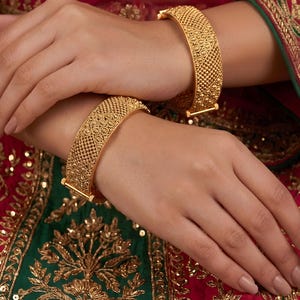 May include: Two gold bangles featuring a textured, intricate lattice pattern with small raised dots. The bracelets have a wide, flat band design with a polished finish, showcasing detailed metalwork across the entire surface of each piece.