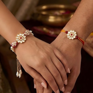 May include: Two decorative wristbands featuring central flower-shaped motifs with white stones and red centers. The left band includes red enamel accents with gold detailing and dangling pearl-like beads, while the right band uses a simple yellow cord.