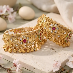 May include: A pair of gold-toned bangles featuring intricate filigree metalwork, embedded with clear white stones and central circular red stones. The edges are lined with small white beads and dangling gold charms along the bottom rim.