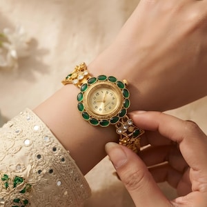 May include: A gold-tone analogue wristwatch featuring a round face with a champagne dial and black numerals. The bezel is encircled by oval-shaped green cabochon stones. The ornate metal bracelet includes intricate filigree details and clear crystals.