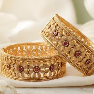 May include: Two gold-toned bangles featuring an intricate openwork floral pattern. Each flower is centered with a small, round, deep red stone. The edges of the bangles have a textured, beaded finish, and the metal has a matte, brushed appearance.