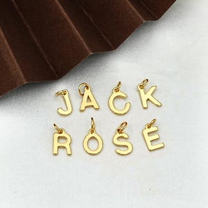 Puede incluir: Amuletos dorados con letras que deletrean los nombres JACK y ROSE. Cada letra tiene un pequeño lazo en la parte superior para sujetarlo a un collar o pulsera. Los amuletos se muestran sobre una superficie blanca.