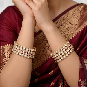 May include: Gold-coloured bracelets, each with multiple rows of clear, round gemstones. The bracelets are worn on the wrists, against a maroon fabric with gold patterns. The jewellery is a statement piece.