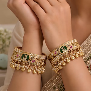 May include: A pair of wide gold-tone bangles featuring a central circular green enamel inlay, accented with small pink stones and rows of white beads. The lower edge of each bangle is decorated with dangling gold-tone charms and small white pearls.