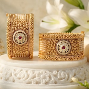 May include: Two wide gold bangles featuring intricate textured metalwork, rows of small white pearls, and a central circular medallion. Each medallion contains a single deep red stone surrounded by clear white stones set in a gold frame.