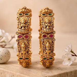 May include: Two gold-toned bangles featuring intricate embossed figures of deities, accented with small oval-shaped green and pink stones and tiny clear crystals. The metal surface has a matte finish with detailed carvings throughout the design.